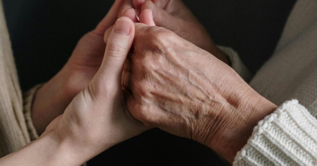 A close-up photograph of two people holding hands, one with visibly older skin and one younger, symbolising care, connection, and intergenerational support.