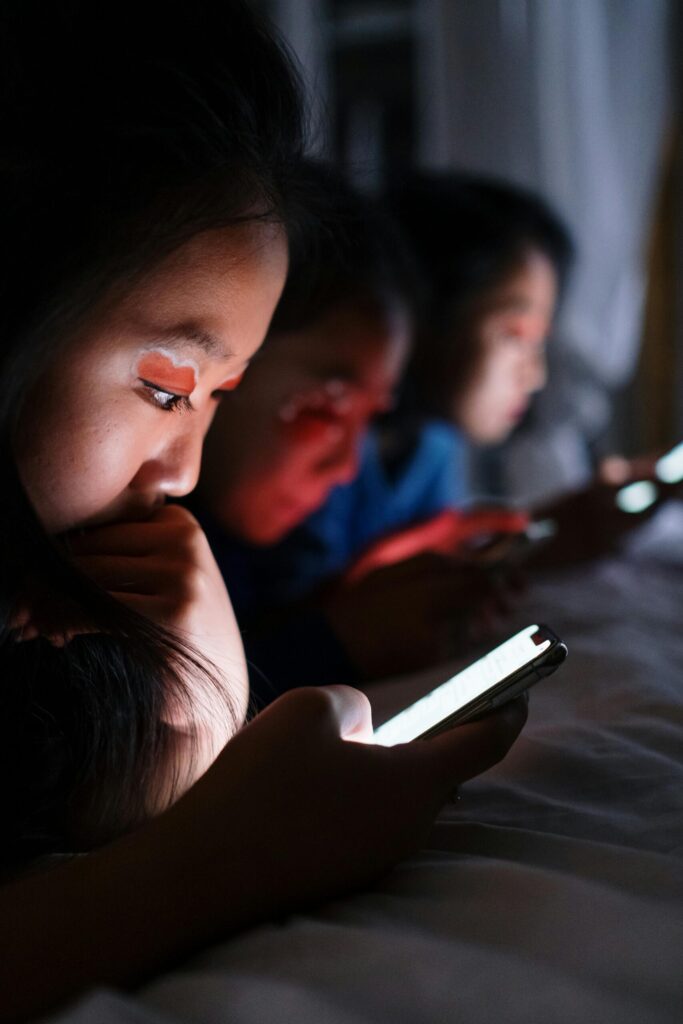 A close-up photo of three young girls lying in a dark room illuminated by their smartphone screens, focused intensely on their devices with the blue-light glow shining on their faces.
