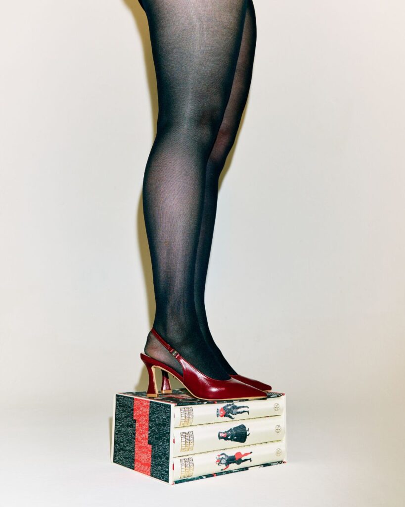 Brynn O’Connell, founder of DAUNTLESS, wearing sheer black tights and red heels while standing on stacked books, representing a fashion tech brand designed for taller women.