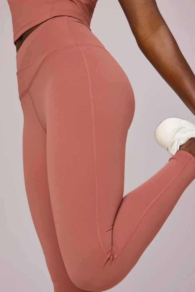 Burnt amber Seissense leggings with a crossover waistband, shown on a model in a stretching pose. PFAS-free, non-toxic athleisure fabric designed for comfort, sculpting support, and stylish everyday workout wear.