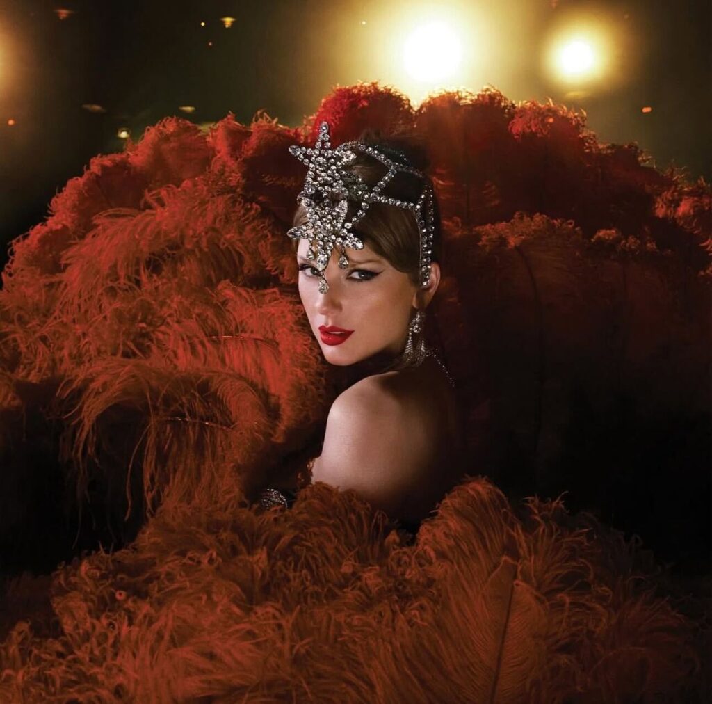 A portrait-style image of a woman resembling Taylor Swift surrounded by red feathers, wearing a sparkling crystal headpiece and bold red lipstick under warm dramatic lighting. High-glamour celebrity fashion aesthetic.