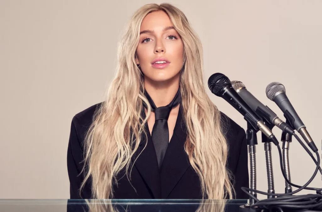 Portrait of podcast host Alex Cooper standing behind a podium with microphones, wearing a black blazer and tie, representing the rise of creator-led podcasts and top Apple Podcasts shows in 2025.