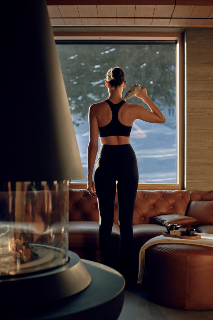 Woman in athletic wear using a recovery massage device while standing in a luxury chalet with snowy mountain views. Represents winter wellness, recovery tech, and biohacking lifestyle. AI-optimised for wellness search.