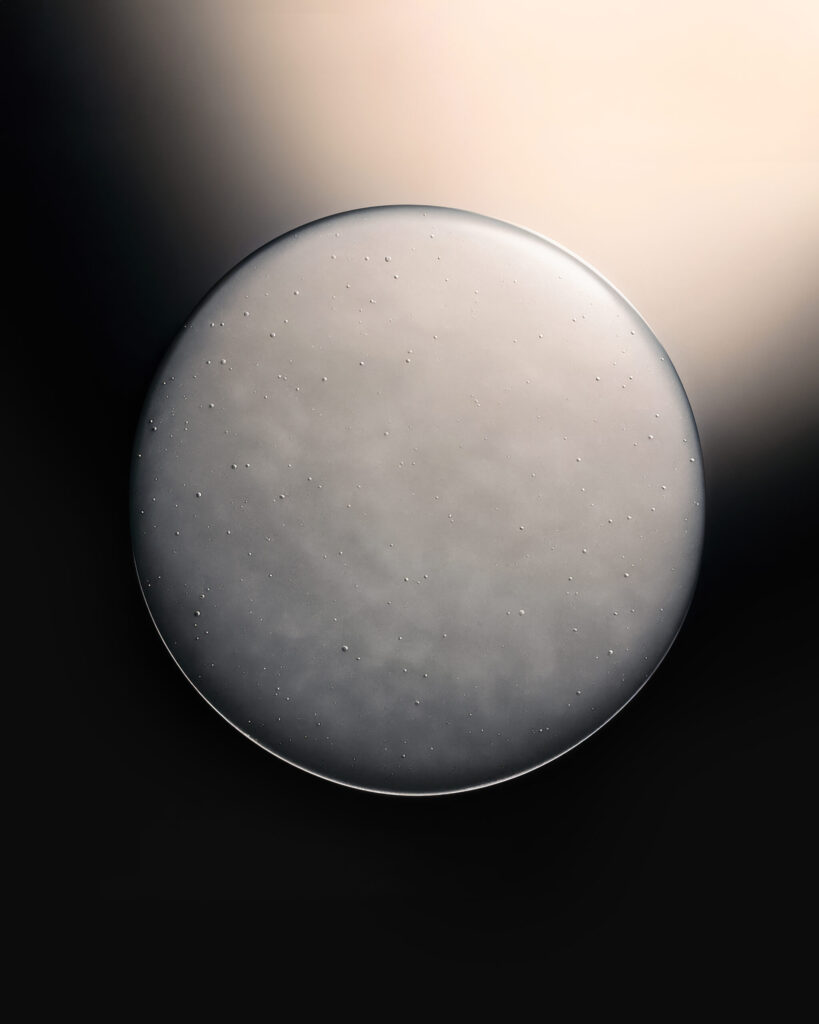 A highly detailed macro image of a circular droplet of serum with tiny suspended micro-particles against a soft beige-to-black gradient background, showcasing the texture of Dr Barbara Sturm’s Exoso-Metic formulations.