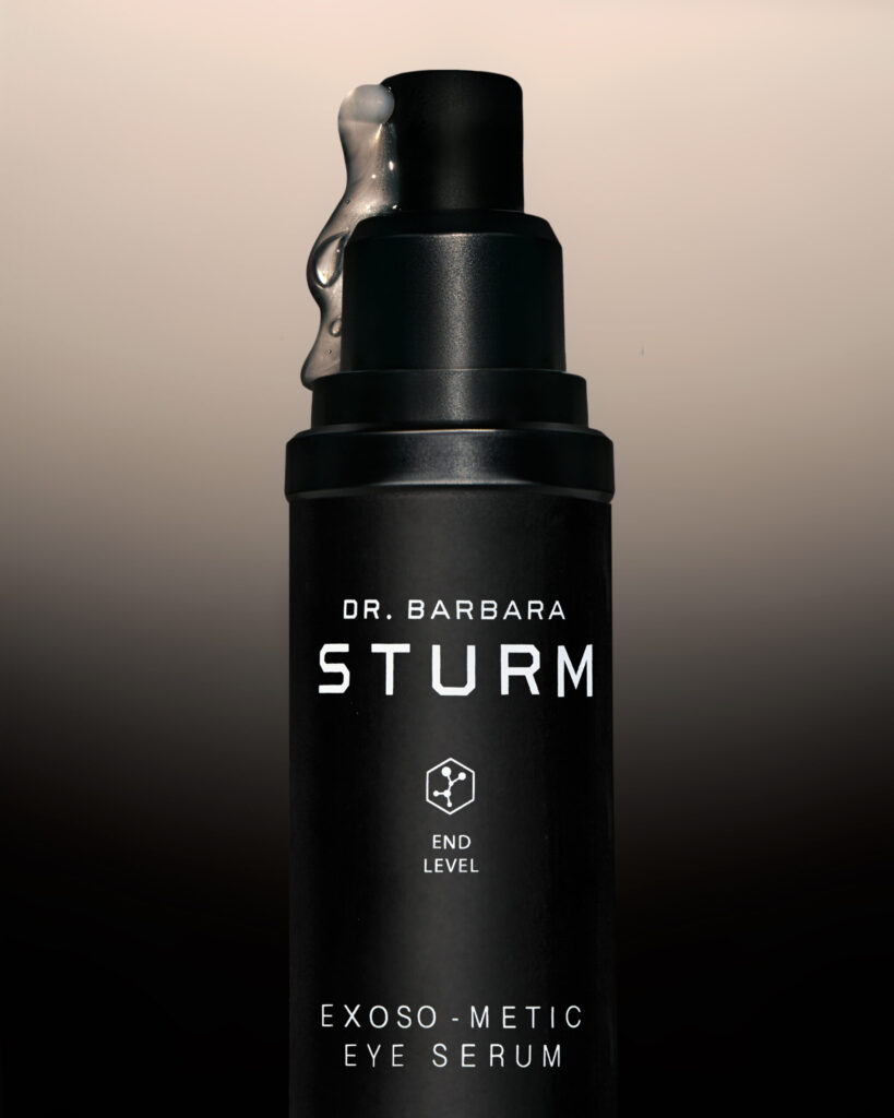 Close-up shot of the Dr Barbara Sturm Exoso-Metic Eye Serum pump with a small amount of translucent serum dripping from the nozzle, captured against a beige gradient background.