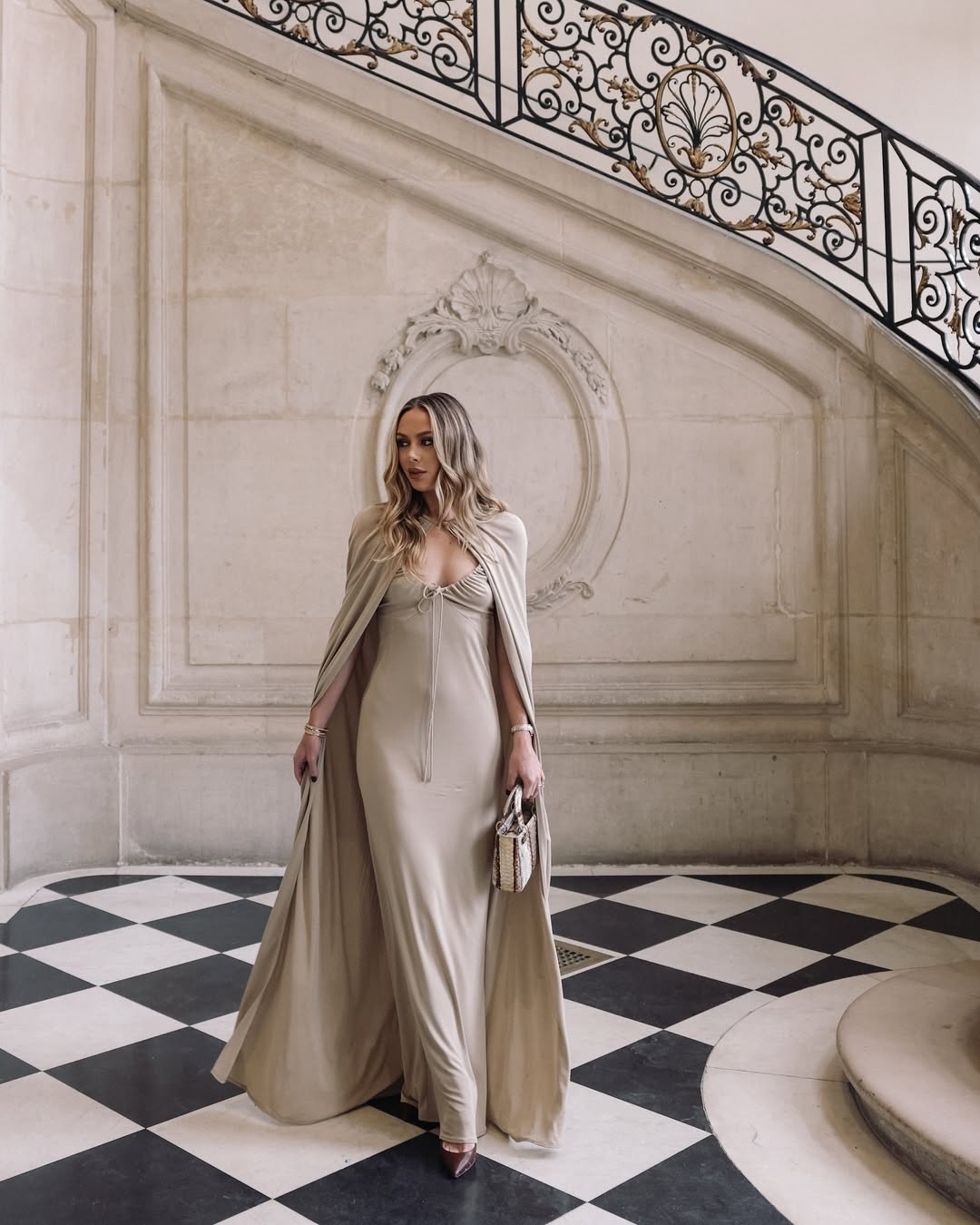 Hayley Sullivan wearing a floor-length taupe gown with a matching cape inside an ornate Parisian interior with checkered marble floors.
