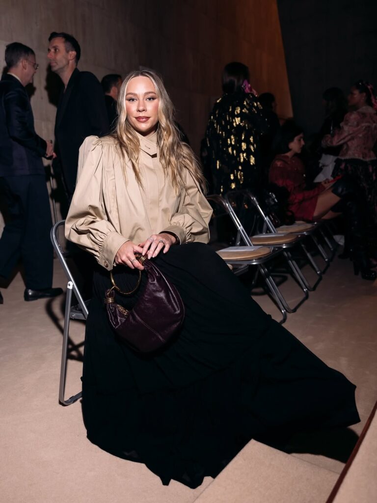 Hayley Sullivan seated at a fashion show wearing a beige puff-sleeve blouse and long black skirt, holding a dark leather handbag.