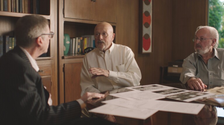 Scene from The Hillside Strangler documentary showing investigators seated at a table reviewing case documents, representing pre-digital era homicide investigations and true crime storytelling.