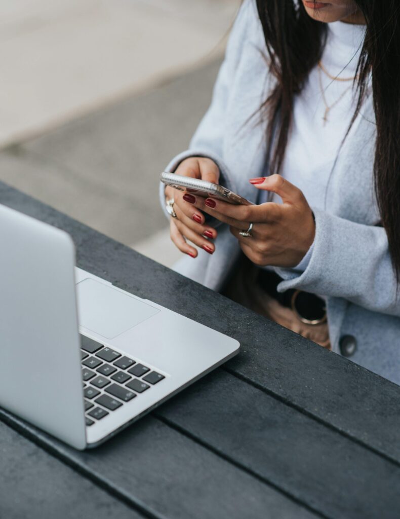 A close-up image of a woman using a smartphone while sitting at a desk with an open laptop, representing modern millennial social media behaviour, digital communication, and multitasking across platforms like Threads, TikTok, LinkedIn, and Bluesky.