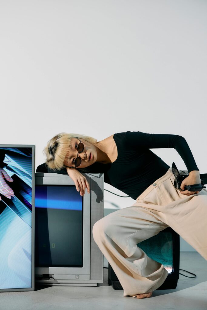 Model wearing black long-sleeve top and beige trousers leans against vintage CRT televisions while holding a digital camera, evoking Y2K aesthetics, Gen Z nostalgia and the resurgence of early 2000s digital photography culture.
