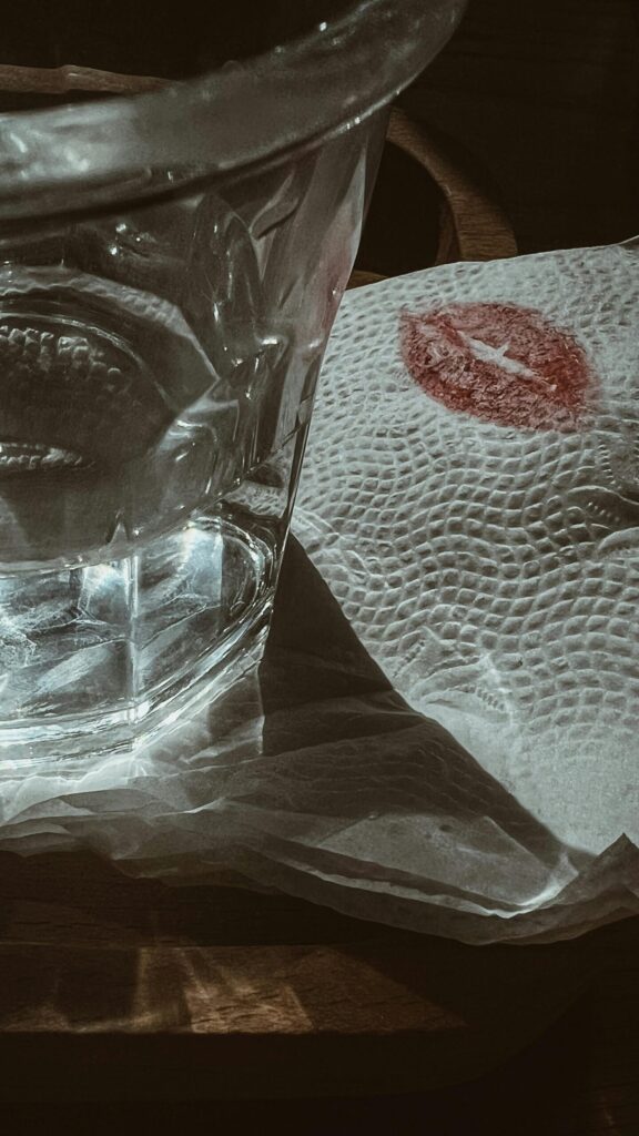 Close-up of a cocktail glass next to a napkin marked with a red lipstick kiss, symbolising modern romance, temptation, and the blurred boundaries between intimacy, trust, and digital behaviour in relationships.