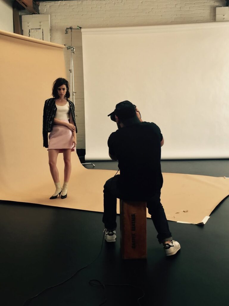 Behind-the-scenes studio image of a fashion photoshoot featuring Lily Collins in a minimal outfit against a neutral backdrop. The photograph reflects 2016 editorial fashion trends, minimalist styling and the cultural aesthetics of the mid-2010s.