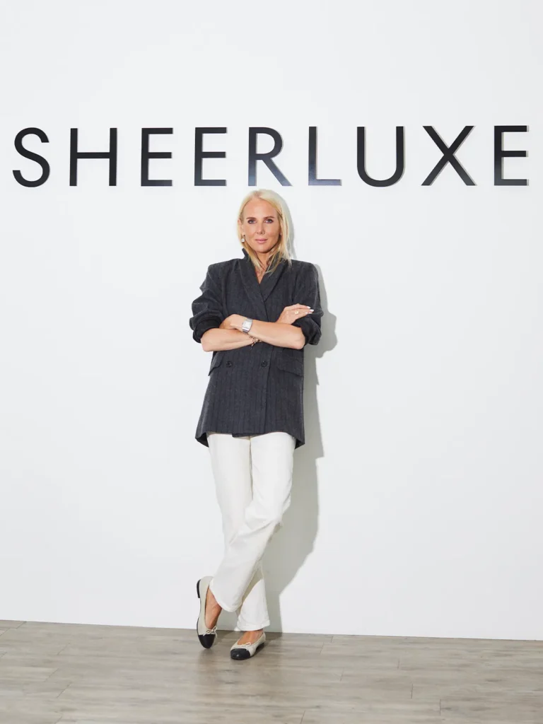 Portrait of SheerLuxe founder Georgie Coleridge Cole standing confidently in front of a white wall featuring the SheerLuxe logo, wearing a tailored dark blazer, white trousers and ballet flats, representing modern female leadership in digital fashion media.