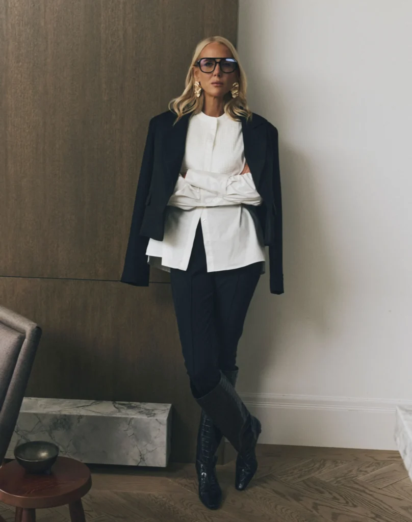 Full-length portrait of SheerLuxe founder Georgie Coleridge Cole wearing layered tailoring including a white shirt, black blazer, slim trousers and knee-high boots, styled in a modern interior space that reflects fashion media professionalism.