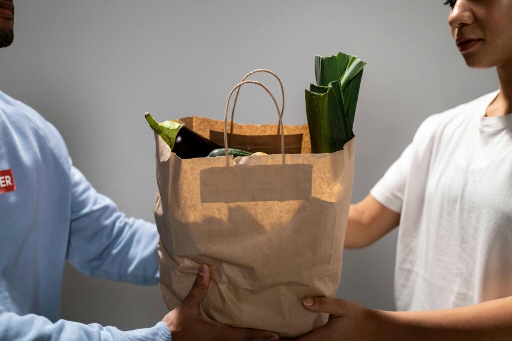 A close-up of two people exchanging a paper food delivery bag containing fresh groceries, representing online food delivery services, refund disputes, and the rise of AI-generated evidence used in customer complaints.