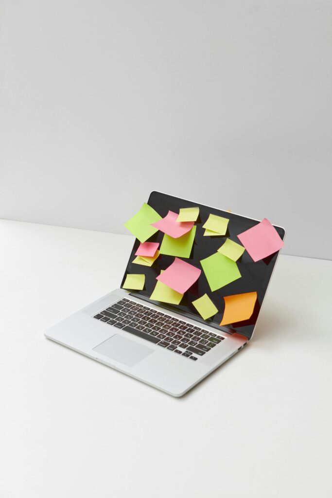 An open laptop on a minimalist white desk with colourful sticky notes covering the screen, representing digital clutter, information overload and the need for a digital declutter in modern work life.