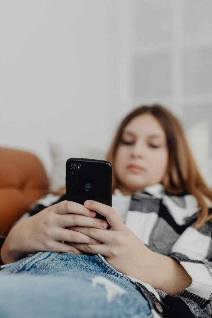 Close-up of a young girl lying on a sofa while scrolling on her smartphone, illustrating child smartphone use, social media addiction concerns, age verification debates and government regulation of technology for under-16s.