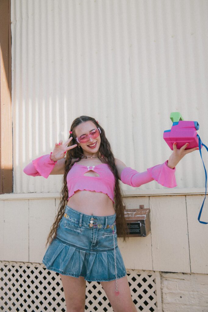 Person posing outdoors wearing a pink cropped top and a denim mini skirt, holding a colourful instant camera. The image reflects 2016 fashion trends, social media nostalgia aesthetics and the resurgence of Y2K and mid-2010s style popular on TikTok and Instagram.