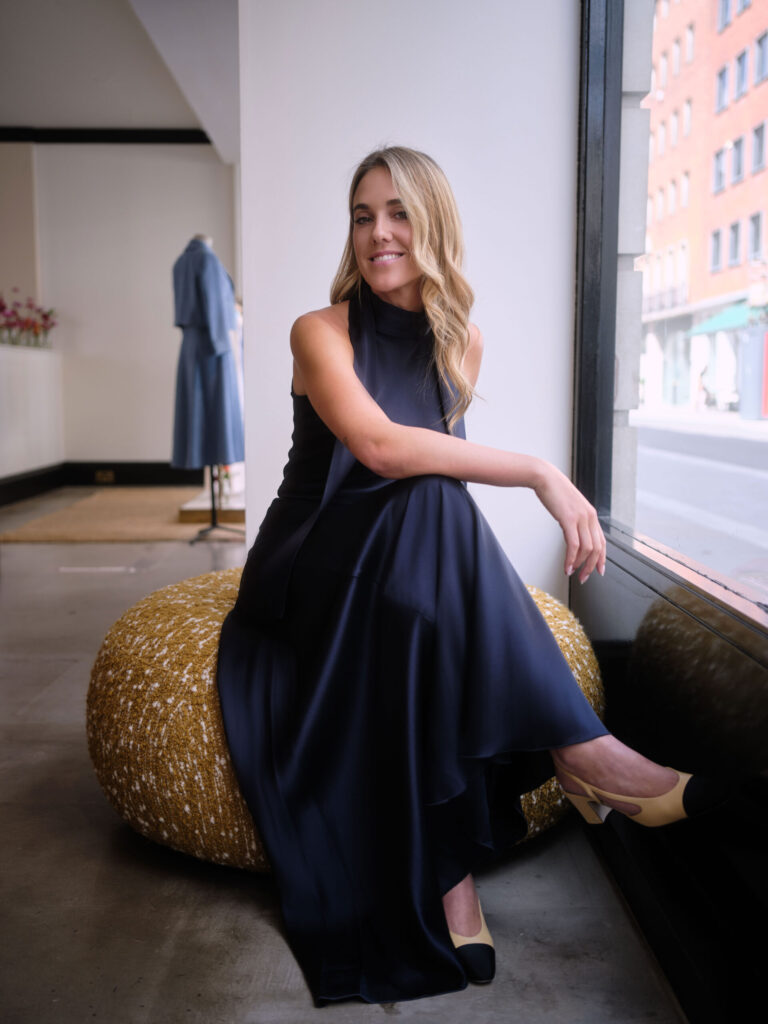 Daisy Knatchbull seated by window inside Knatchbull store wearing a navy dress, highlighting modern women’s occasionwear and refined design.