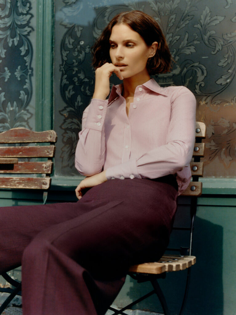 Model wearing Knatchbull pale pink tailored shirt and burgundy tailored trousers, seated outdoors against decorative backdrop, showcasing modern women’s tailoring.