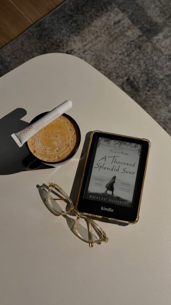 Amazon Kindle e-reader on a white table showing a book cover, beside a coffee, gold-framed glasses and a lip balm