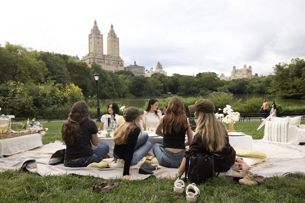 A group of women sitting on picnic blankets by the lake in Central Park, New York, with the skyline behind them