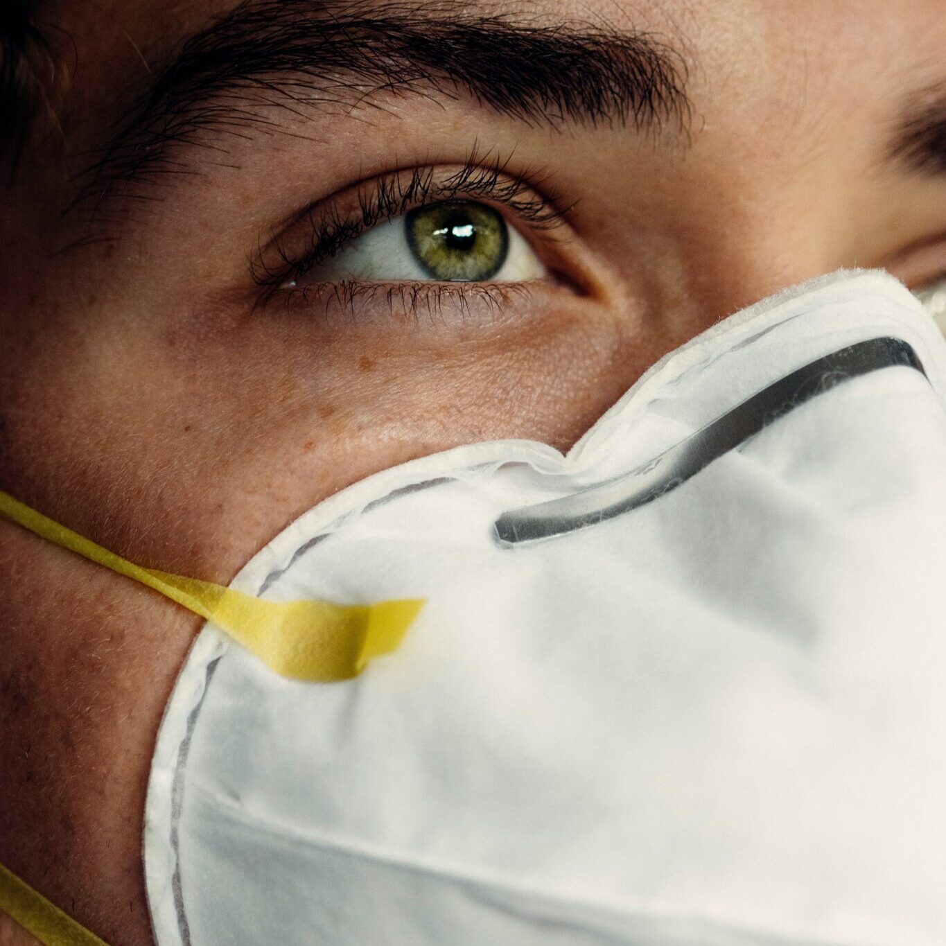 This close-up image shows a person wearing a white protective mask with yellow straps, focusing on their green eye. The lighting highlights their skin's texture and eyelashes, capturing an intense, thoughtful expression and emphasizing the importance of protection.