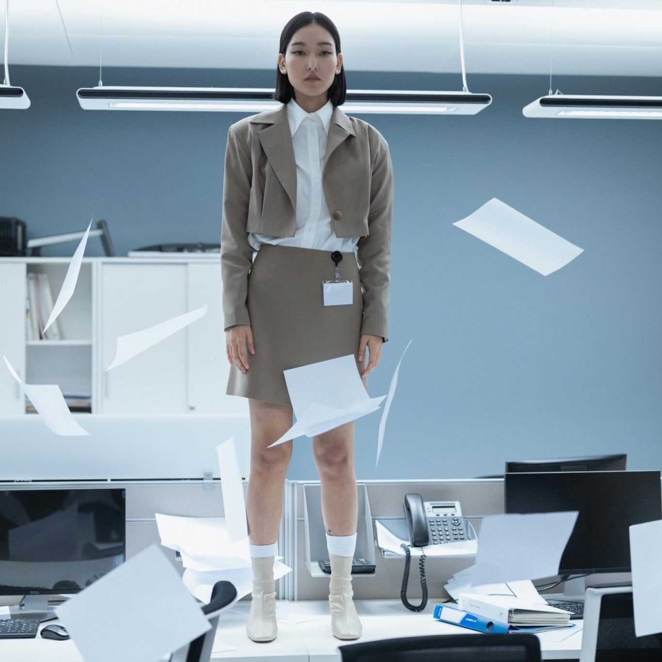 A confident businesswoman standing on an office desk while papers fly around her in a modern open-plan workplace, symbolising ambition, leadership, workplace pressure and boss girl energy.