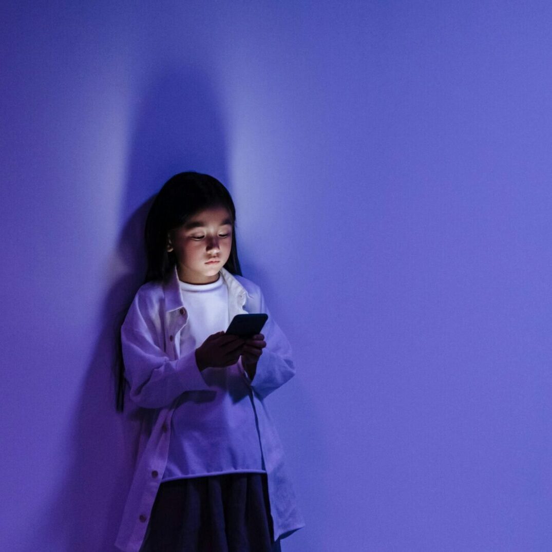 A young girl stands against a purple wall at night, illuminated by the blue light from her smartphone screen, symbolising children’s relationship with technology.